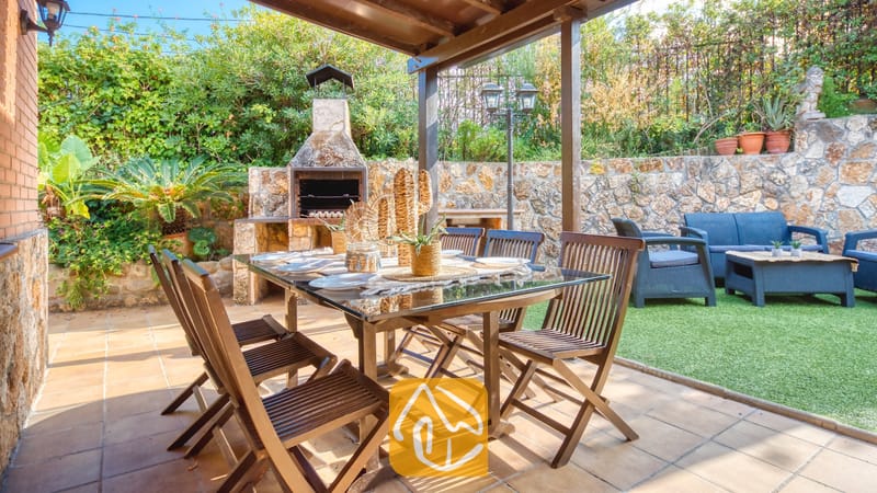 Patio extérieur couvert de la Villa Alba, doté d'une table en bois surmontée d'une plaque en verre, de chaises pliantes et d'un barbecue en pierre intégré en arrière-plan.