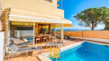 Piscine et terrasse de la Villa Santa Cristina, agrémentées de tomettes, de bains de soleil gris et d'un espace repas en bois ombragé par un store jaune, offrant une vue imprenable sur la mer.