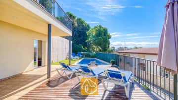 Terrasse en bois ensoleillée à la Villa Mauri, agrémentée de transats bleus, avec une piscine en contrebas et un patio couvert sous un ciel bleu éclatant