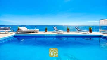 Piscine d'un bleu éclatant à la Villa Infinity avec des transats blancs et des plantes en pot sur la terrasse carrelée au bord de l'eau, surplombant la mer