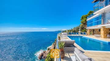 Piscine à débordement à flanc de falaise à la Villa Infinity, avec des transats bleus et de vastes vues sur la mer sous un ciel d'un bleu éclatant