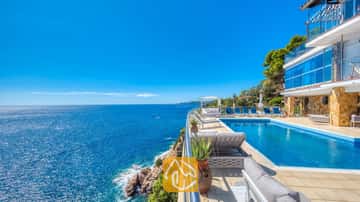 Piscine à débordement à flanc de falaise à la Villa Infinity, avec des transats bleus et de vastes vues sur la mer sous un ciel d'un bleu éclatant