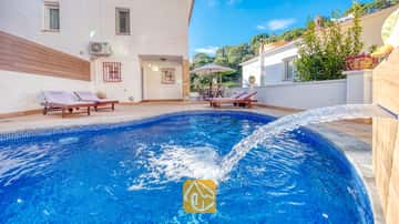 Piscine incurvée en carrelage bleu dotée d'une fontaine à la Villa Blanca, agrémentée de chaises longues en bois sur la terrasse pavée, adossée à un mur extérieur blanc.