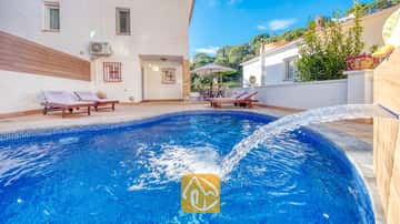 Piscine incurvée en carrelage bleu dotée d'une fontaine à la Villa Blanca, agrémentée de chaises longues en bois sur la terrasse pavée, adossée à un mur extérieur blanc.