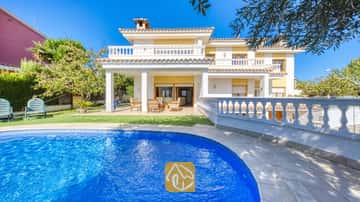 Piscine aux carreaux de mosaïque bleue en premier plan, avec la Villa Baileys arborant une façade blanche, des murs jaunes et des balcons carrelés sous un ciel dégagé.