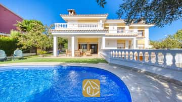 Piscine aux carreaux de mosaïque bleue en premier plan, avec la Villa Baileys arborant une façade blanche, des murs jaunes et des balcons carrelés sous un ciel dégagé.