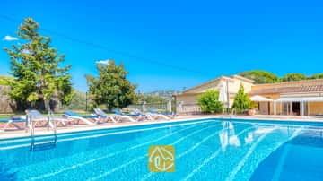 Piscine extérieure à la Villa Miro, agrémentée de chaises longues bleues et blanches sur une terrasse en terre cuite. Des arbres verdoyants et une bâtisse jaune se détachent sous un ciel bleu azur.