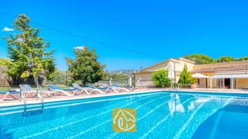 Piscine extérieure à la Villa Miro, agrémentée de chaises longues bleues et blanches sur une terrasse en terre cuite. Des arbres verdoyants et une bâtisse jaune se détachent sous un ciel bleu azur.