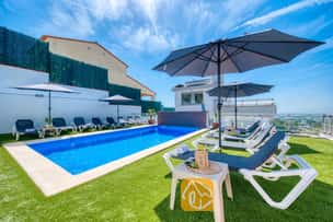 Piscine privée à la Villa Sandy Beach, entourée de gazon synthétique, de chaises longues grises à structure blanche et de grands parasols sous un ciel bleu azur.