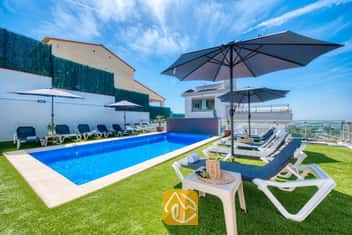 Piscine privée à la Villa Sandy Beach, entourée de gazon synthétique, de chaises longues grises à structure blanche et de grands parasols sous un ciel bleu azur.