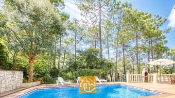Piscine privée à la Villa Esmee avec des chaises longues blanches sur la terrasse carrelée, entourée de grands pins et dotée d'un barbecue en pierre.