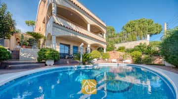 Piscine aux formes courbes avec carrelage en mosaïque, située devant la façade à plusieurs étages de la Villa Grace, qui arbore des arches en pierre et des balcons sous un ciel d'un bleu limpide.
