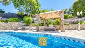 Piscine privée à la Villa Lorena avec margelles en carrelage bleu, tonnelle en tissu beige surplombant le salon de jardin, murs de soutènement en pierre et végétation environnante.