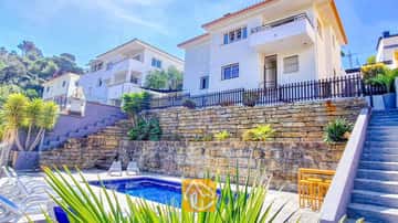 Vue extérieure de la Villa Fransisca présentant une piscine rectangulaire adossée à des murets de pierre en terrasses, sous un bâtiment blanc à plusieurs étages et un ciel bleu.