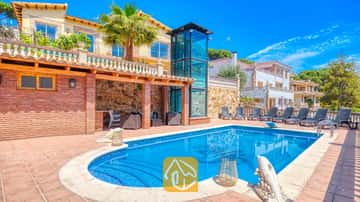 Piscine et terrasse privées à la Villa Dolce Vita, dotée d'une cage d'ascenseur en verre, de murs en briques et de plusieurs chaises longues sous un ciel bleu.