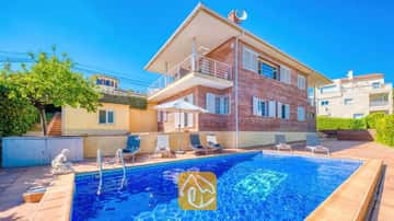 Vue extérieure de la Villa Beyoncé mettant en valeur une piscine rectangulaire en carrelage bleu, des chaises longues et un bâtiment en briques de deux étages sous un ciel bleu éclatant