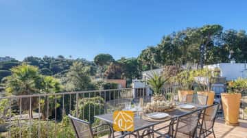 Terrasse ensoleillée à la Casa AdoRa dotée d'une table à manger en métal dressée avec des assiettes et des verres blancs, surplombant des arbres verdoyants sous un ciel bleu.