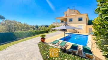 Vue extérieure de la Villa Holiday montrant la maison jaune à deux étages, la piscine dotée d'une mosaïque en forme de dauphin, ainsi que les transats verts installés sous un parasol gris.