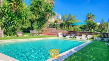 Piscine extérieure à la Villa Summertime entourée d'une pelouse verdoyante, de chaises longues, d'un parasol bleu et de palmiers tropicaux sous un ciel d'un bleu éclatant