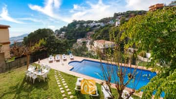 La Villa Marysol dispose d'une piscine extérieure entourée de bains de soleil blancs, d'une table à manger sur la pelouse et d'un village à flanc de colline en arrière-plan.