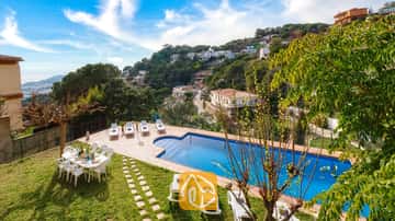 La Villa Marysol dispose d'une piscine extérieure entourée de bains de soleil blancs, d'une table à manger sur la pelouse et d'un village à flanc de colline en arrière-plan.