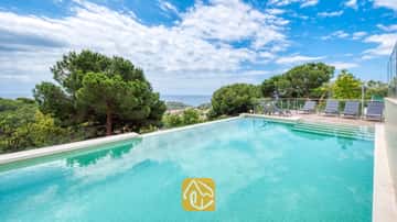Piscine privée à la Villa BellaVista, agrémentée de carreaux turquoise, d'une terrasse ensoleillée clôturée de verre avec des chaises longues grises, offrant une vue imprenable sur la mer et les pins.