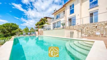 Grande piscine extérieure avec escalier incurvé à la Villa BellaVista. L'extérieur en pierre est agrémenté de terrasses aux garde-corps en verre, d'une plage en bois et de bains de soleil gris, le tout entouré d'une végétation luxuriante.