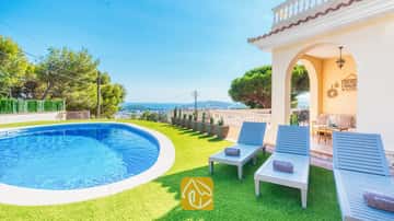 Piscine privée et transats en bois bleu sur gazon synthétique à la Villa Ibiza, avec vue sur la mer au loin et les arbres.