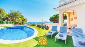 Piscine privée et transats en bois bleu sur gazon synthétique à la Villa Ibiza, avec vue sur la mer au loin et les arbres.