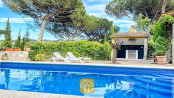 Piscine privée aux rebords carrelés de bleu et chaises longues blanches sur la terrasse de la villa Iako, agrémentée d'un espace barbecue extérieur et de grands the pins en arrière-plan.