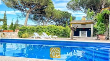 Piscine privée aux rebords carrelés de bleu et chaises longues blanches sur la terrasse de la villa Iako, agrémentée d'un espace barbecue extérieur et de grands the pins en arrière-plan.