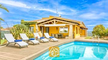 Piscine privée avec terrasse en bois et transats blancs à la Villa Soleia en Espagne, caractérisée par une villa extérieure jaune et un ciel bleu.