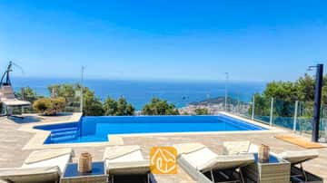 Piscine privée et terrasse carrelée à la Villa VistaMar, dotée de transats blancs, de garde-corps en verre et d'une vue lointaine sur la côte sous un ciel d'un bleu éclatant.