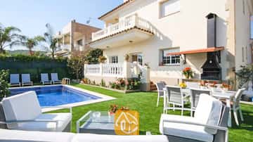 Vue extérieure de la Villa Mercedes Fenals présentant une piscine, une pelouse, un salon de jardin blanc et un barbecue extérieur intégré sur fond de façade couleur crème.